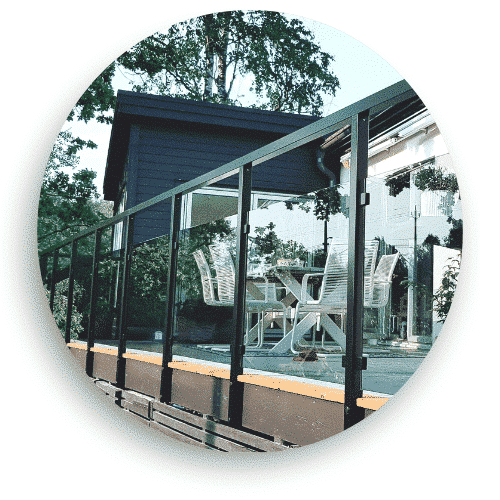 Glasräcke med svarta aluminiumprofiler och genomskinliga glasskivor, monterat på en altan med trädäck. Räcket har en tunn överliggare och skapar en avgränsning runt uteplatsen. I bakgrunden syns en utemöbelgrupp och en fasad med speglande glaspartier.