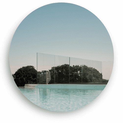 Genomskinligt glasräcke utan stolpar monterat vid en poolkant. De stora glasskivorna hålls på plats av en diskret bottenprofil, vilket ger en obruten utsikt över landskapet i bakgrunden. Vattnet reflekterar himlen och skapar en harmonisk miljö.