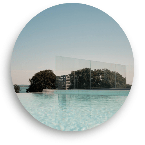 Pare-vent en verre transparent autour d’une piscine extérieure, avec vue dégagée sur arbres et mer en arrière-plan.