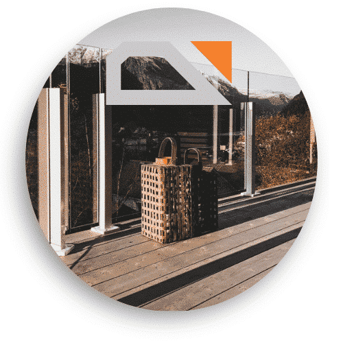 Glasräcke med vita stolpar och klämfästen monterat på en altan med trägolv. Räckets transparenta glaspartier ramar in utsikten över berg och dalgång. Två dekorativa lyktor står på trallen.