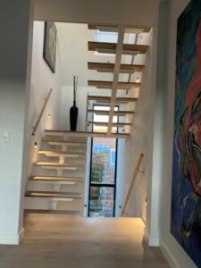 Escalier suspendu en bois clair avec limon central blanc, rampe murale et grande fenêtre pour une lumière naturelle abondante.