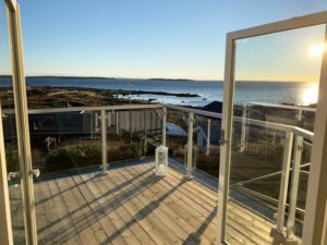 Garde-corps en verre avec structure blanche sur balcon en bois, surplombant la mer au coucher du soleil.