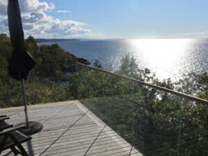 Rambarde de terrasse en verre avec main courante en bois, vue dégagée sur lac scintillant sous ciel bleu.